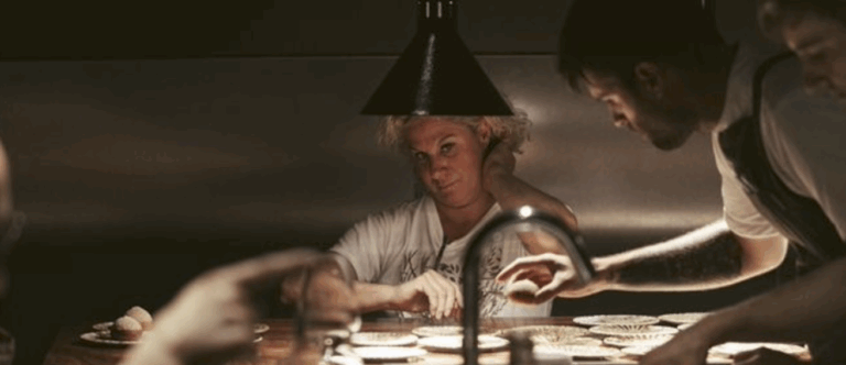 Sous une lumière tamisée qui éclaire le passe comme une scène de théâtre, la cheffe Ana Roš observe attentivement Samuel Chastenay, concentré sur le dressage des assiettes. Son regard perçant trahit à la fois l’exigence et la confiance qu’elle accorde à son équipe. Samuel, absorbé par le geste, travaille avec précision, entouré de collègues également à l'œuvre. L’ambiance est tendue mais maîtrisée : chaque mouvement compte dans cette cuisine trois étoiles Michelin.