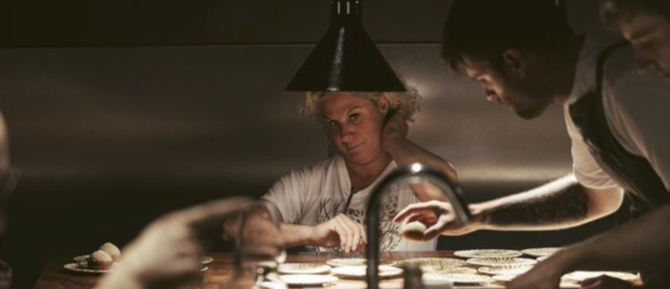 Sous une lumière tamisée qui éclaire le passe comme une scène de théâtre, la cheffe Ana Roš observe attentivement Samuel Chastenay, concentré sur le dressage des assiettes. Son regard perçant trahit à la fois l’exigence et la confiance qu’elle accorde à son équipe. Samuel, absorbé par le geste, travaille avec précision, entouré de collègues également à l'œuvre. L’ambiance est tendue mais maîtrisée : chaque mouvement compte dans cette cuisine trois étoiles Michelin.