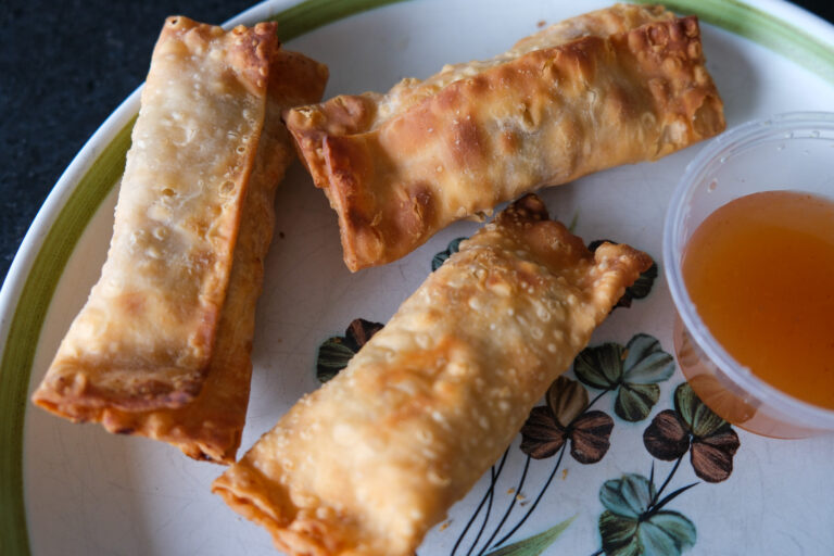 Trois egg rolls dorés, disposés sur une assiette blanche à motifs floraux avec des teintes vertes et brunes. Leur pâte est croustillante et bien frite, avec une texture légèrement bosselée qui suggère un extérieur croquant. À côté des egg rolls, un petit contenant de sauce orangée est visible, une sauce aigre-douce prête à accompagner ces bouchées savoureuses.