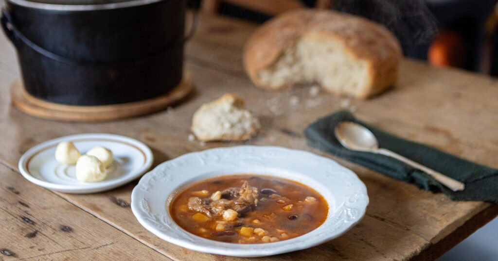 Un plat de sagamité servi dans un bol blanc délicatement orné. La sagamité, une soupe traditionnelle autochtone, est composée ici de morceaux de viande, probablement de gibier, accompagnés de maïs et de haricots. La couleur riche du bouillon laisse entrevoir un mélange savoureux et réconfortant. À côté du bol, une cuillère repose sur une serviette en tissu vert foncé, ajoutant une touche de simplicité et d'authenticité rustique.