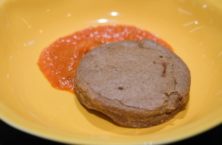 Ce plat, signé Christian Têtedoie, met en valeur le haricot viande, une légumineuse locale et ancienne, dans une présentation à la fois rustique et élégante. La galette, d’une teinte brune naturelle, évoque un produit fait maison, travaillé avec soin. Elle est accompagnée d’une purée de tomates ou d’une sauce rouge veloutée qui ajoute une touche de couleur vibrante et un contraste visuel dans l’assiette. Ce mariage de simplicité et de technique culinaire illustre parfaitement la philosophie du chef : sublimer des ingrédients humbles tout en offrant une expérience gustative sophistiquée.