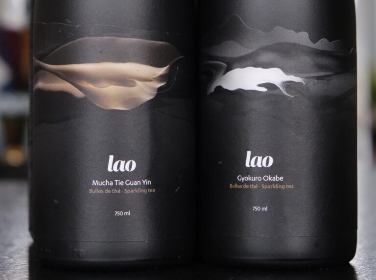 La photo montre deux bouteilles élégantes de bulles de thé produites par l'entreprise Lao. Les bouteilles, en verre noir mat, arborent un design minimaliste et sophistiqué. Chaque étiquette présente un motif abstrait évoquant des mouvements fluides, en tons dorés et argentés.