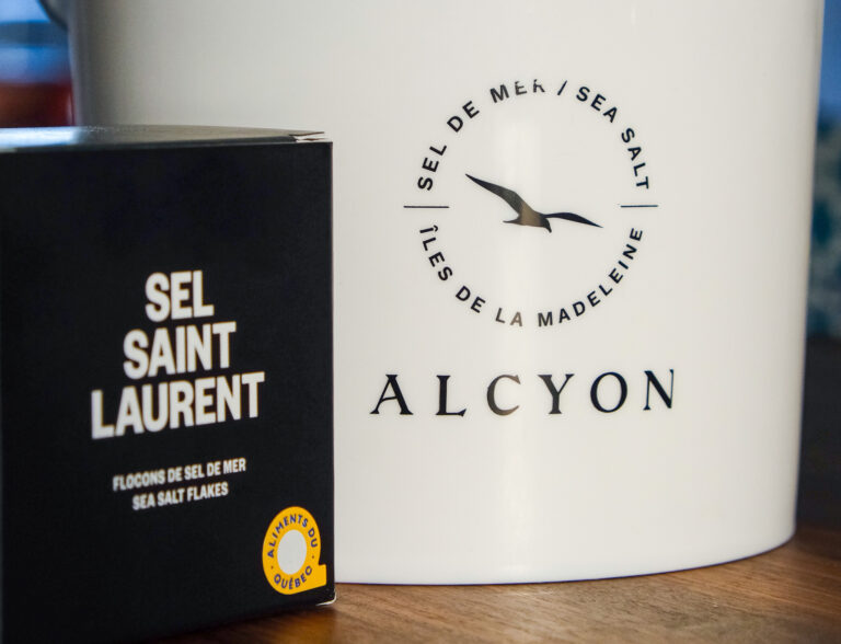 L'image présente une chaudière blanche et une boîte noire. Le pot est plus grand et sert probablement de contenant. Il est décoré d'un cercle avec l'inscription "SEL DE MER / SEA SALT" en haut et "ILES DE LA MADELEINE" en bas. Au centre du cercle, on peut voir le dessin stylisé d'un oiseau en vol. La boîte noire, plus petite, est posée à côté du pot. Elle porte l'inscription "SEL SAINT LAURENT" en grandes lettres blanches et "FLOCONS DE SEL DE MER / SEA SALT FLAKES" en plus petites lettres. On peut également apercevoir un petit logo représentant un poisson, suggérant ainsi un produit alimentaire.