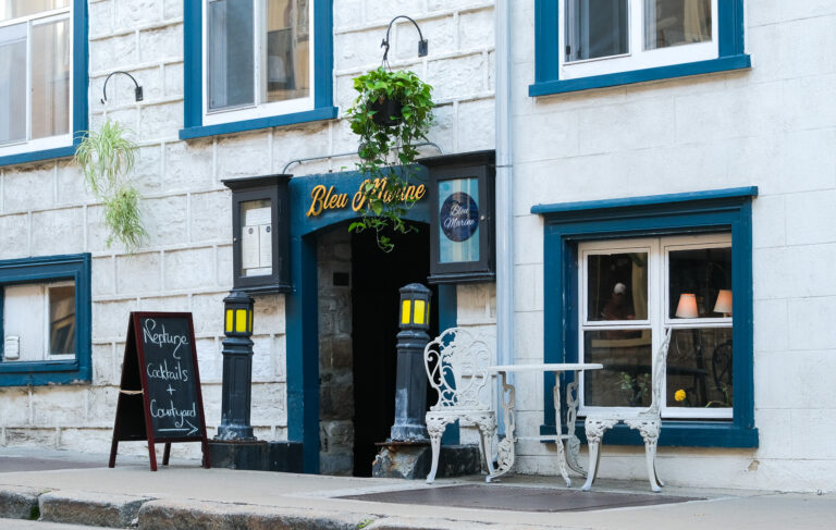 Cette photo représente une façade de bâtiment accueillante et charmante, avec une enseigne indiquant "Bleu Marine" en lettres cursives jaunes. Le mur est en pierre claire, contrastant élégamment avec des encadrements de fenêtres peints en bleu. Une ardoise sur le trottoir annonce "Neptune Cocktails + Courtyard", suggérant une expérience conviviale, possiblement dans une cour intérieure. Une table et des chaises blanches au design vintage ajoutent une touche romantique et rétro à l'ambiance. Une plante suspendue au-dessus de l'entrée et des lanternes extérieures complètent l'atmosphère chaleureuse et accueillante de l'endroit.