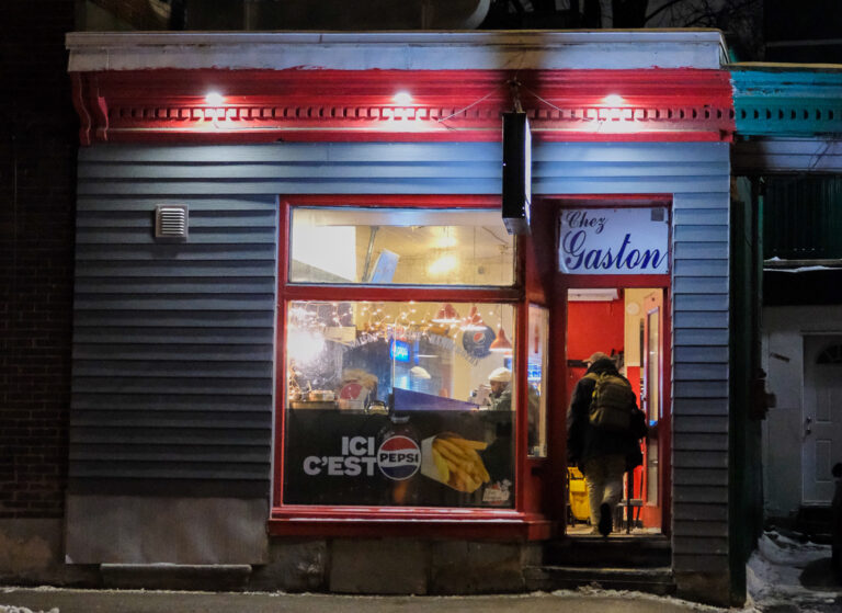 La photo montre la façade du casse-croûte Chez Gaston, situé dans le quartier Saint-Roch à Québec, de nuit. L’enseigne bleue et blanche avec l’écriture cursive « Chez Gaston » est bien visible au-dessus de la porte d’entrée rouge. L’extérieur du bâtiment est peint en bleu-gris avec des moulures rouges et une touche de turquoise sur la corniche. À travers la grande fenêtre, on peut apercevoir l’intérieur du casse-croûte, illuminé et animé, avec des cuisiniers en action derrière le comptoir. Une affiche sur la vitre indique « ICI C’EST PEPSI », accompagnée d’une image de frites.