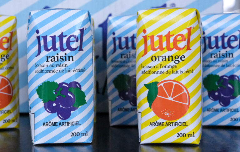 Les boîtes sont clairement identifiées avec le logo « Jutel » en grandes lettres blanches sur le devant. On peut également lire « Jutel » plus discrètement sur le côté des boîtes. Il est précisé qu'il s'agit d'une « boisson au raisin » ou d'une « boisson à l'orange », avec la mention « additionnée de lait écrémé » en dessous. Les boîtes de jus Jutel sont dans un contexte qui évoque la production ou la distribution en grande quantité. Le design simple et les couleurs vives mettent en avant le produit et son identité de marque.
