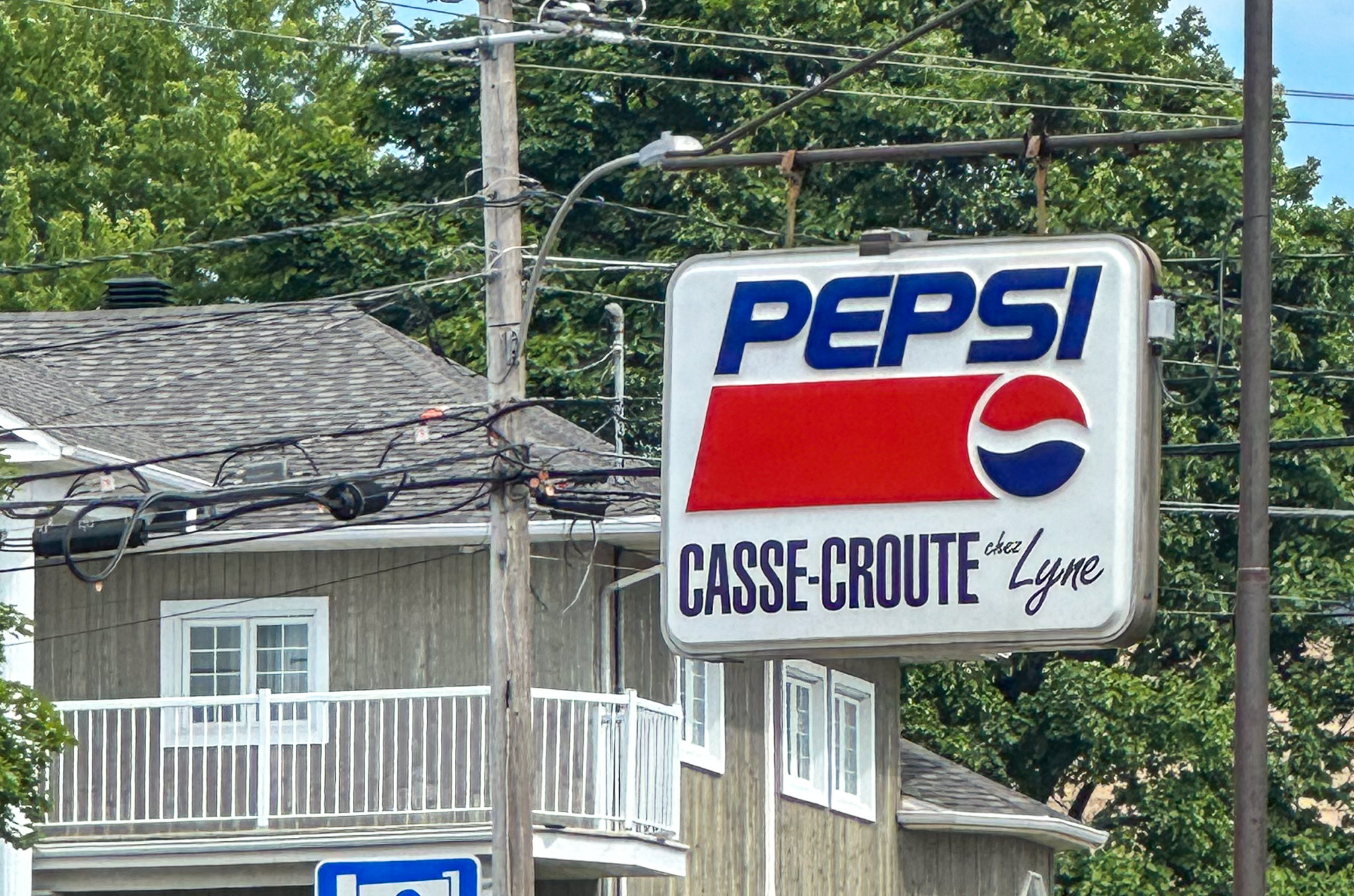 Une enseigne rétro bien typique des casse-croûtes québécois. L’enseigne affiche le logo classique de Pepsi, avec ses couleurs rouge, blanc et bleu, accompagné du nom de l’établissement Casse-Croûte chez Lyne, écrit dans une police manuscrite. Le panneau est suspendu à un vieux poteau électrique en bois, encadré de câbles qui accentuent le caractère brut et authentique du lieu. En arrière-plan, on voit une maison ou un bâtiment résidentiel en bois, avec un balcon blanc, ce qui suggère un environnement de village ou de petite ville typique du Québec. La verdure abondante autour ajoute à l’ambiance estivale et décontractée.