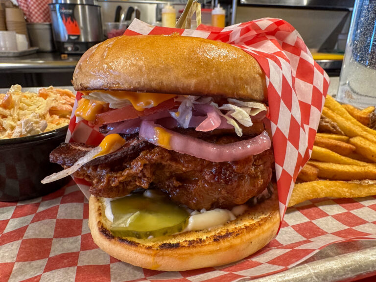 Ce burger ultra-gourmand de La Baraque à frites combine un **poulet frit croustillant**, du **bacon fumé**, une **sauce Buffalo relevée**, des **oignons marinés**, du **fromage fondant**, de la laitue et des tomates, le tout dans un **pain brioché grillé** garni de mayo et de cornichons. Servi avec des **frites dorées** et une **salade de chou crémeuse**, il promet une explosion de saveurs et de textures dans un pur esprit du cuisine réconfort.