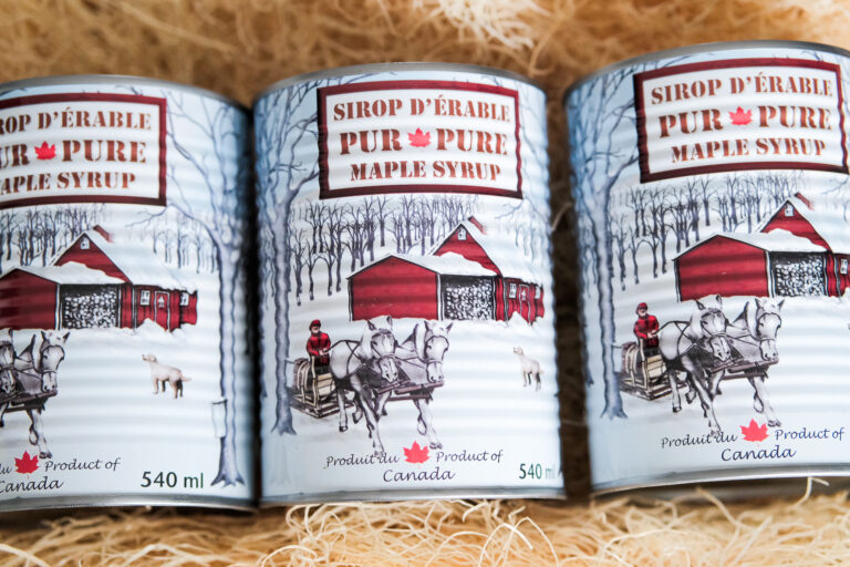 La photo montre trois conserves de **sirop d'érable pur**, un produit emblématique du Canada. Chaque boîte de 540 ml arbore une illustration hivernale traditionnelle avec une cabane à sucre rouge entourée d'érables, des chevaux tirant une carriole et une personne vêtue de rouge, évoquant l'authenticité et le savoir-faire artisanal du sirop d'érable. L'étiquette, bilingue en français et en anglais, met en avant l'origine canadienne du produit avec une feuille d'érable rouge. L'arrière-plan en paille ajoute une touche rustique, renforçant l’image artisanale et locale du produit.