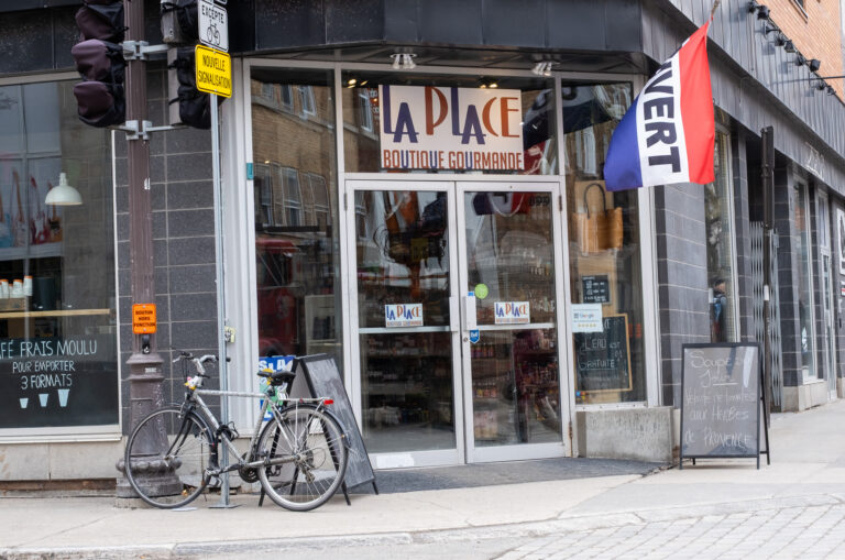 Sur cette photo, on découvre La Place – Boutique Gourmande, une charmante épicerie de quartier située au coin d'une rue à Québec. La façade moderne en briques grises est agrémentée d’une grande vitrine qui laisse entrevoir les étagères bien garnies de produits. Au-dessus de la porte vitrée double, une enseigne colorée indique le nom du commerce, et un drapeau rouge, blanc et bleu portant la mention "OUVERT" flotte pour accueillir les passants. À droite de l’entrée, un petit tableau noir annonce la soupe du jour : Velouté de tomates aux herbes de Provence, écrite à la craie, ajoutant une touche artisanale et conviviale. Une affiche signale aussi que l’eau est gratuite, soulignant l’aspect accueillant et accessible de la boutique. Sur le trottoir, un vélo est attaché à un poteau de signalisation, accentuant l’ambiance urbaine et locale. À gauche, on aperçoit une autre vitrine avec une affiche pour du café frais moulu à emporter.