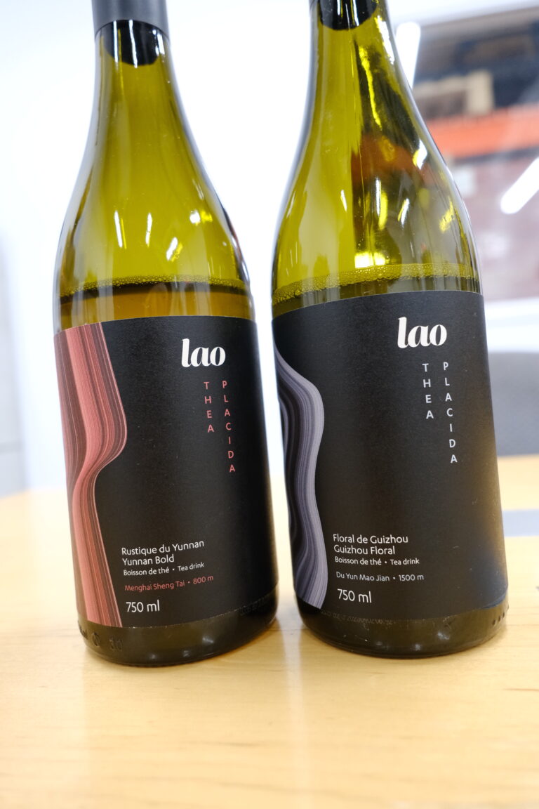 Sur cette photo, on voit deux élégantes bouteilles en verre de 750 ml aux lignes rappelant celles du vin, posées côte à côte sur une table en bois pâle. Elles arborent des étiquettes noires sobres et raffinées, ornées d’un motif ondulé vertical coloré — rouge pour la bouteille de gauche, violet-gris pour celle de droite. La marque lao est inscrite en lettres blanches en haut de chaque étiquette, suivie du nom de la gamme Thea Placida, qui évoque calme et sérénité. La bouteille de gauche contient la cuvée Rustique du Yunnan (Yunnan Bold), une boisson de thé à base de Menghai Sheng Tai. Celle de droite, quant à elle, propose une version plus délicate et florale : Floral de Guizhou (Guizhou Floral), issue du Du Yun Mao Jian.
