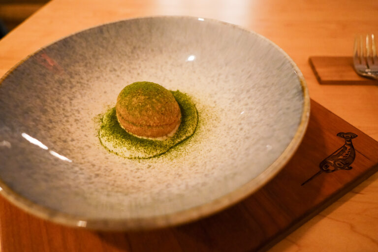 Sur cette photo, on découvre une élégante réinterprétation du pani puri, servie dans un bol aux teintes minérales posé sur une planche de bois gravée d’un narval stylisé — clin d’œil au nom du restaurant. Au centre de l’assiette trône une sphère dorée, croustillante, délicatement posée sur un nuage de crème blanche, le tout saupoudré d’une fine poudre verte aux accents boréaux. Un plat à la fois intrigant et raffiné, qui évoque l’équilibre entre nature sauvage et influences voyageuses, signature du chef Norman St-Pierre.