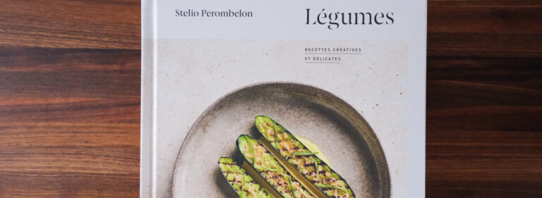 La photo montre la couverture du livre Légumes – Recettes créatives et délicates du chef Stelio Perombelon, posée sur une table en bois foncé aux reflets chaleureux. Le design du livre est sobre et élégant : en haut, le nom de l’auteur est inscrit en noir sur fond blanc, à gauche, et le titre Légumes apparaît à droite, dans une typographie raffinée. En dessous, on lit Recettes créatives et délicates, en lettres capitales espacées. L’image centrale présente une assiette en céramique texturée, aux tons naturels, dans laquelle reposent trois courgettes coupées en deux, garnies d’un mélange finement haché, dressées sur un lit de sauce jaune. L’ensemble évoque à la fois la précision du geste culinaire et l’esthétique épurée propre à la cuisine du chef.