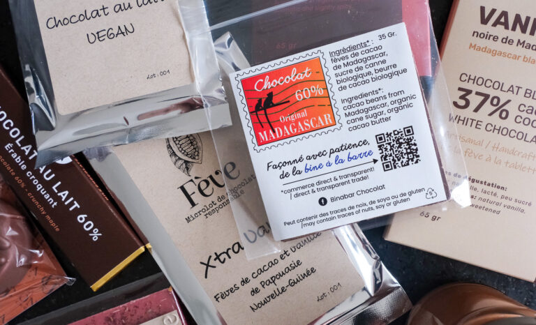 La photo présente un bel assortiment de tablettes de chocolat artisanal fabriquées de la fève à la tablette, issues de micro-lots et soigneusement emballées. On y voit clairement trois marques : Fève, Binabar et ONOA, toutes originaires de la région de Québec. Au centre, une tablette de chocolat Binabar 60 % cacao "Original Madagascar" attire l’attention avec son étiquette colorée. On y lit la liste d’ingrédients simples : fèves de cacao de Madagascar, sucre de canne biologique et beurre de cacao biologique. Une mention souligne que le chocolat est « façonné avec patience, de la fève à la barre » et que l’entreprise mise sur un commerce direct et transparent. Autour, on distingue une tablette de la chocolatière Fève, notée "XtraVan" à base de fèves de cacao et de vanille de Papouasie-Nouvelle-Guinée, ainsi qu’un chocolat au lait végan. Une autre tablette d’ONOA, au chocolat blanc 37 % cacao à la vanille noire de Madagascar, complète l’ensemble. L’accent est mis sur la qualité des ingrédients, la traçabilité et l’approche artisanale, qui transparaissent autant dans les choix typographiques que dans la clarté des étiquettes.