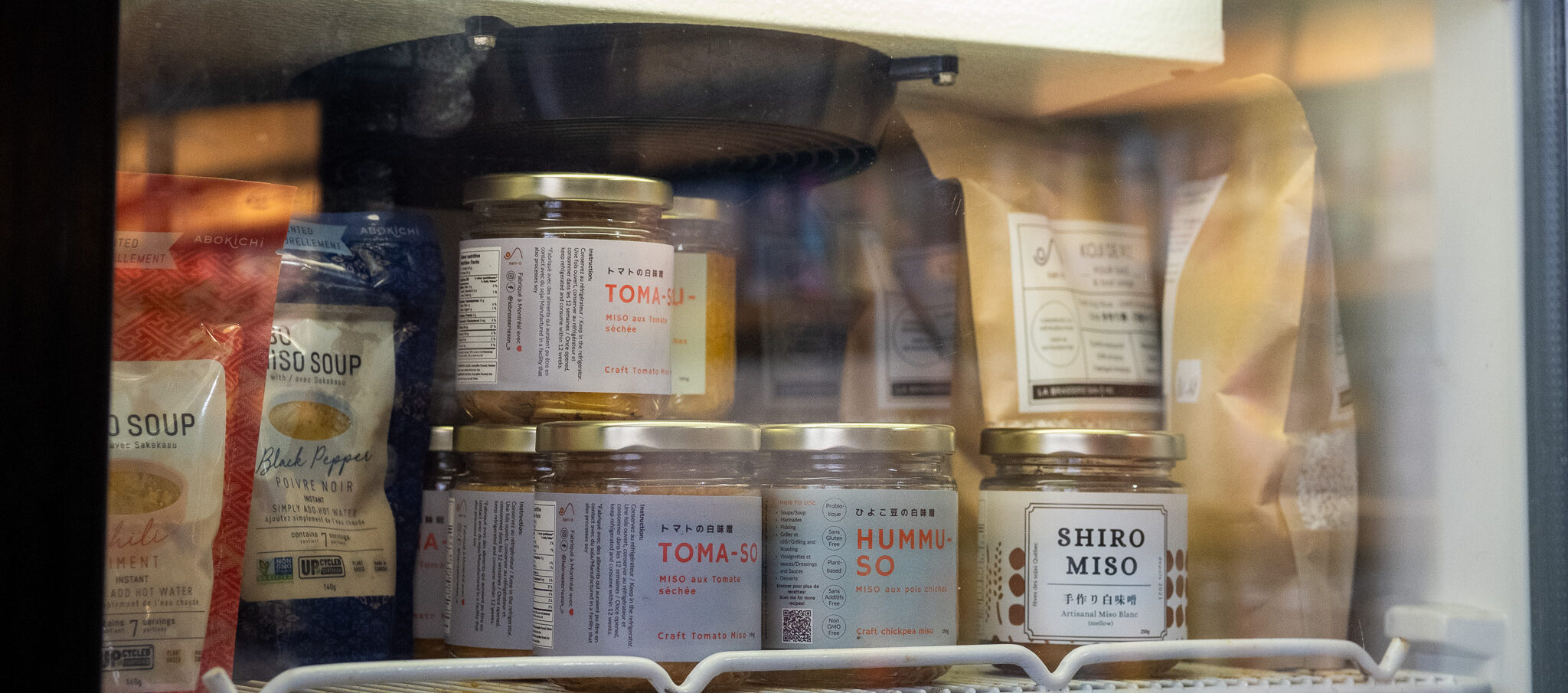 La photo montre l’intérieur d’un réfrigérateur étiqueté « Produits fermentés » au marqueur noir, dans une écriture artisanale peinte à la main sur une bande blanche. Sur les étagères, on aperçoit une belle sélection de produits fermentés, principalement d'inspiration japonaise. Des pots de Toma-su (miso aux tomates séchées), Hummu-so (miso aux pois chiches) et Shiro miso (miso blanc artisanal) sont bien en évidence à l’avant. À gauche, on distingue des sachets de soupe miso instantanée de la marque Abokichi, tandis qu’à l’arrière-plan, des sacs kraft non identifiés complètent l’offre. L’ensemble évoque un espace dédié à des produits soigneusement sélectionnés, mettant en valeur le savoir-faire artisanal et les bienfaits de la fermentation.