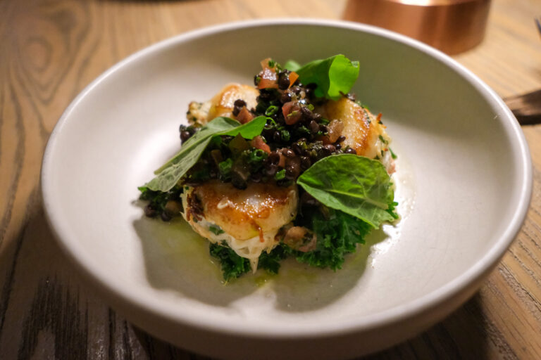 Sur la photo, on découvre une assiette élégante et soigneusement dressée mettant en vedette un pétoncle poêlé à la perfection, saisi jusqu’à obtenir une fine croûte dorée. Le mollusque est farci d’un mélange crémeux au crabe des neiges, déposé sur un lit de verdures poêlées — possiblement du chou kale ou du Tokyo bekana. Le tout est surmonté d’une garniture généreuse de lentilles beluga brillantes, relevées de fines herbes ciselées et de dés de tomate, apportant fraîcheur, texture et une touche d’acidité. Quelques feuilles vertes fraîches viennent couronner l’ensemble, ajoutant à la fois croquant, couleur et vivacité visuelle.