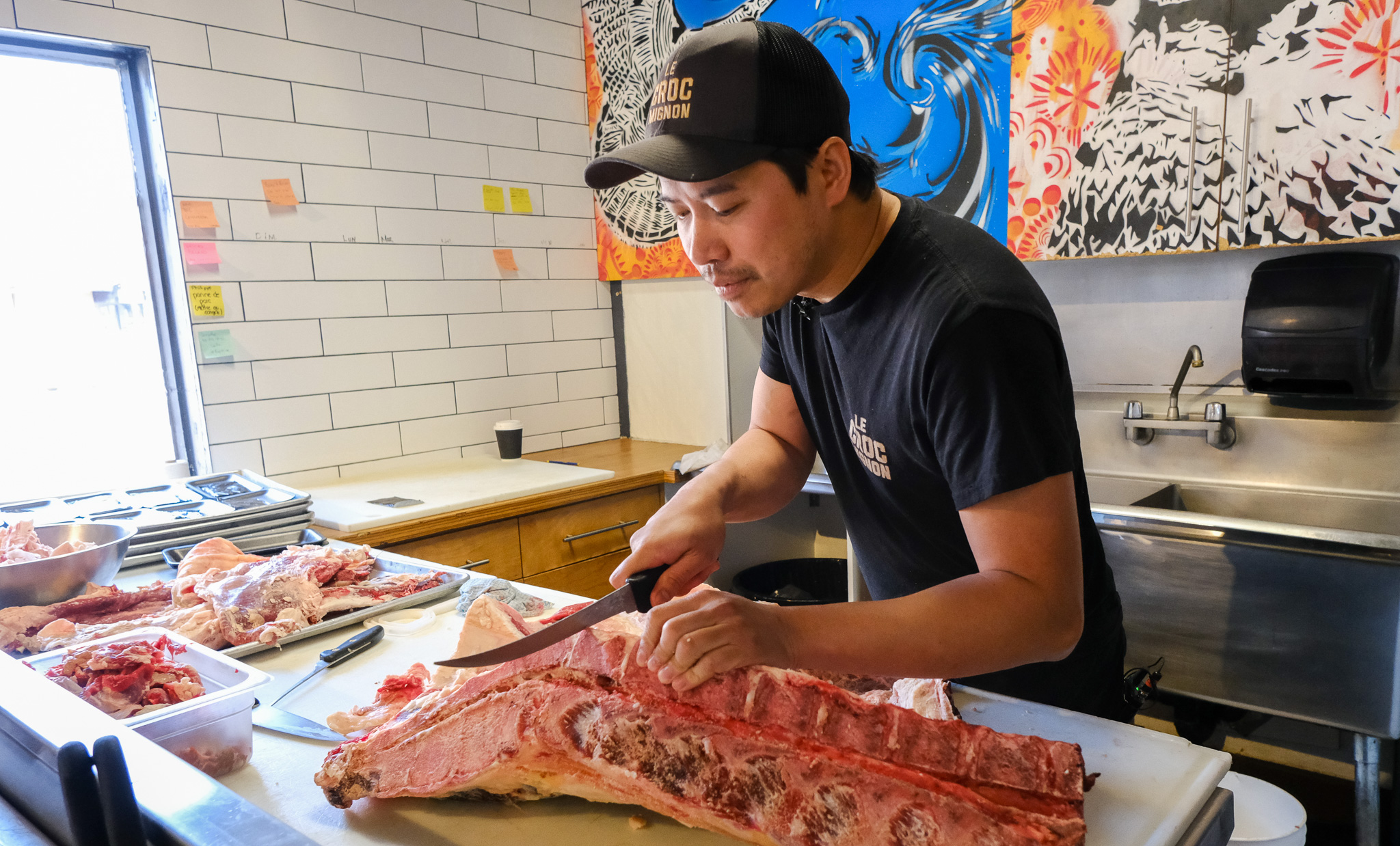Sur cette photo, Sothea Deng, propriétaire de la boucherie Croc Mignon, est concentré sur la découpe méticuleuse d’une côte de bœuf impressionnante. Armé de son couteau de boucher, il travaille directement à partir de la carcasse, dans un espace lumineux et épuré où chaque geste est précis et réfléchi. Derrière lui, des œuvres colorées ajoutent une touche d’humanité et de personnalité à ce lieu de travail artisanal. Cette image illustre parfaitement l’essence de son métier : une boucherie de proximité où la technique rencontre le respect de l’animal et du produit brut.