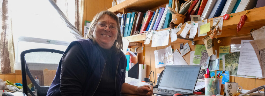 Sur cette photo, on découvre Patricia Gallant, jardinière en cheffe des Jardins de Métis, assise à son bureau dans un petit espace de travail chaleureux et débordant de vie. Souriante et visiblement à l’aise, elle incarne la passion et l’expérience. Autour d’elle, les murs sont tapissés de notes manuscrites, d’images, de calendriers et de documents, témoins de ses nombreuses années de service. Son bureau en bois, bien rempli, évoque un lieu de planification autant que de création, où chaque détail compte. L’écran de son ordinateur affiche un tableau de données — sans doute un plan de plantation ou un suivi des semis. Dans ce capharnaüm organisé, tout respire la rigueur, l’amour du métier et une grande humanité.