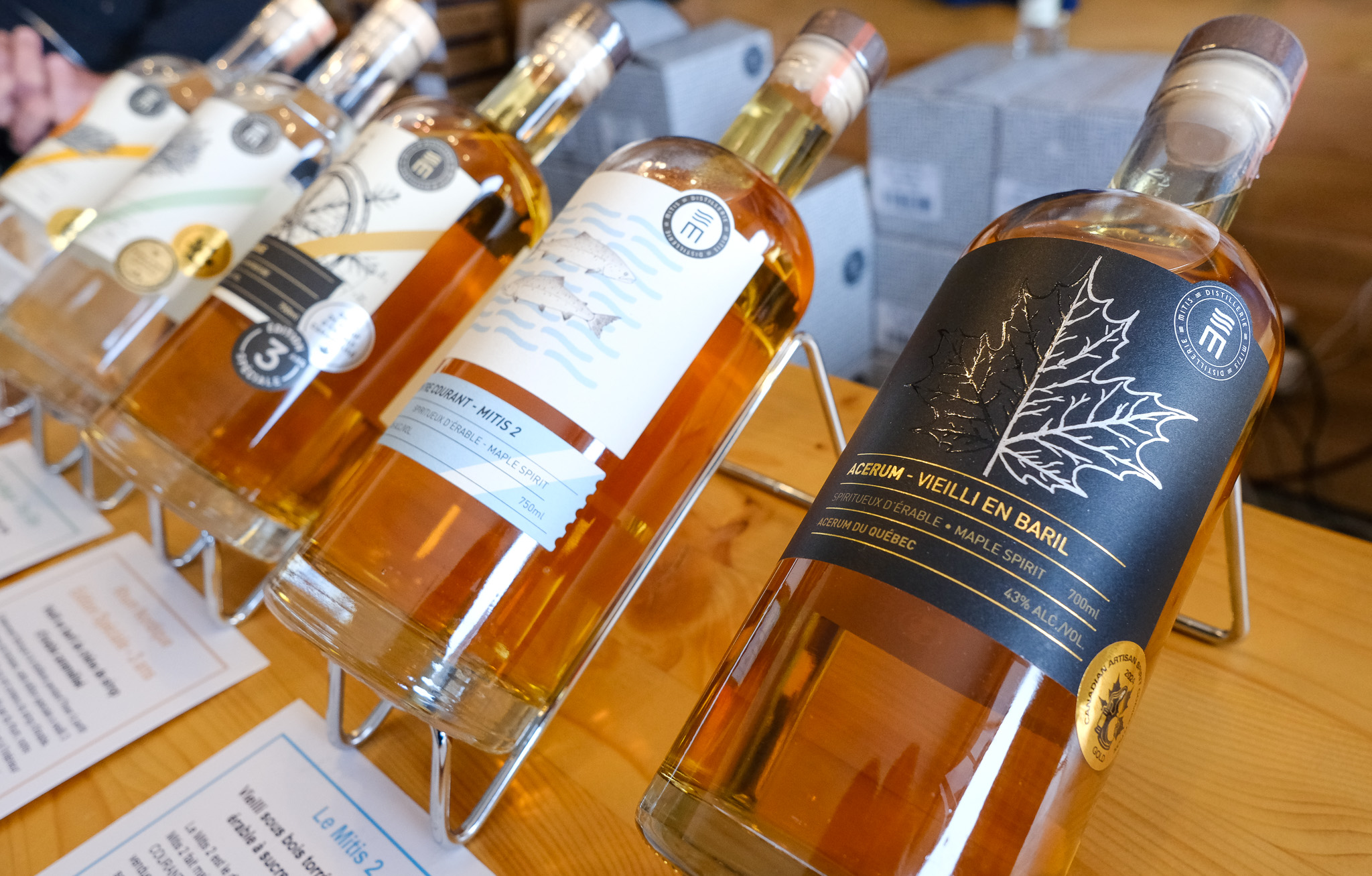 La photo montre une élégante rangée de bouteilles d’acérum artisanales de la Distillerie Mitis, soigneusement alignées sur un présentoir métallique. Les étiquettes raffinées varient de tons clairs à foncés, illustrant différentes cuvées, dont l’« Acérum – Vieilli en baril », au premier plan, identifiable à sa feuille d’érable argentée embossée sur fond noir. Le liquide doré à ambré révèle un vieillissement en fût, et la mention « Spiritueux d’érable – Maple Spirit » affirme clairement l’origine québécoise de ce produit distinctif récemment protégé par une IGP.
