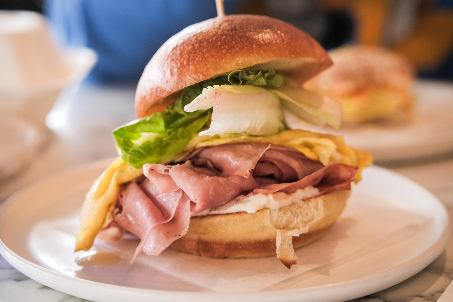 Un sandwich déjeuner généreux et appétissant, servi sur une assiette blanche. Il est composé d’un pain brioché légèrement lustré, bien doré, garni de fines tranches de mortadelle, d’une omelette pliée façon ruban, de feuilles de laitue croquantes et d’une touche de mayonnaise ou de sauce crémeuse. Le tout est maintenu par un petit bâtonnet au centre.