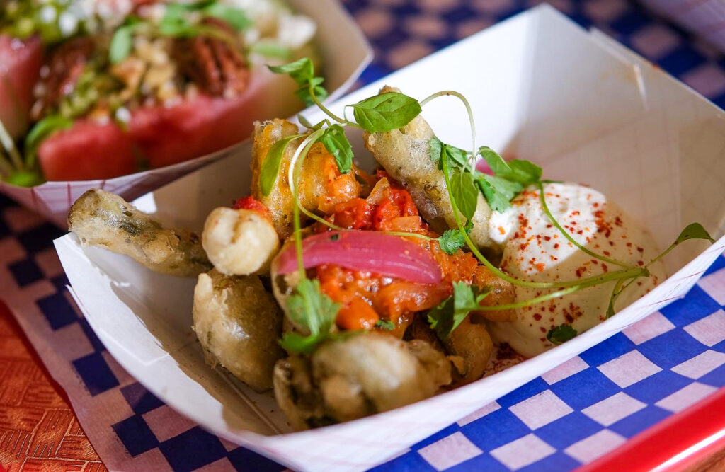 Dans leur barquette rayée façon casse-croûte rétro, les têtes de violon frites volent la vedette. Joliment dorées et croustillantes, elles sont servies avec une généreuse cuillerée de crème fouettée salée, saupoudrée de paprika fumé. Quelques oignons marinés, une salsa de poivrons rouges et des herbes fraîches complètent l’assiette avec panache. Un plat à la fois ludique et savoureux, qui met en valeur un ingrédient emblématique du printemps québécois dans toute sa splendeur.