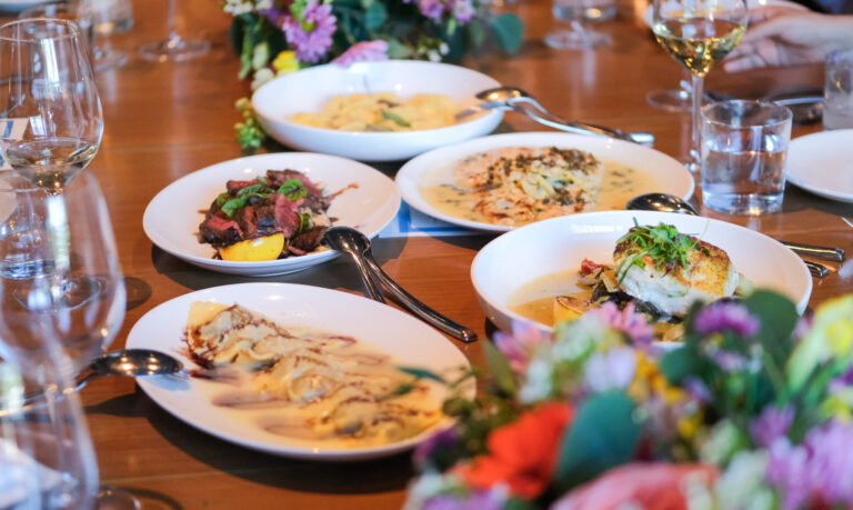 La photo montre une table chaleureusement dressée pour un repas gastronomique, mettant en valeur une variété de plats colorés et soigneusement présentés. On y voit des assiettes de pâtes, un filet de poisson sur un lit de légumes, des tranches de viande grillée garnies de basilic frais, ainsi qu’un plat nappé d’une sauce crémeuse. Des verres de vin blanc, de l’eau et un élégant centre de table fleuri ajoutent une touche festive et raffinée à l’ensemble. L’ambiance évoque un souper entre amis ou un moment de dégustation convivial dans un restaurant haut de gamme.