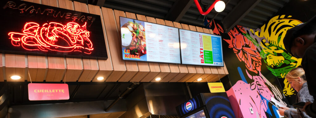 Cette photo capture l’intérieur vibrant et coloré d’un comptoir de restauration rapide asiatique, probablement spécialisé en cuisine cambodgienne, comme l’indique le nom Bati Cantine du Cambodge visible sur le menu numérique. On remarque une ambiance urbaine et moderne, avec un design graphique éclaté : un immense mur illustré de figures stylisées en rose, jaune, vert et bleu, apportant une touche artistique audacieuse à l’espace. Au-dessus du comptoir, deux écrans numériques affichent le menu en détail, avec des sections pour les entrées, plats, à-côtés, breuvages et combos. On y voit aussi une photo appétissante d’un plat nommé « Le Koral ». Un néon rouge accrocheur représentant une figure joyeuse (peut-être un chef ou un personnage symbolique) ajoute une touche rétro et amusante, avec le nom du compte Instagram @cantinebati. Le comptoir est divisé en deux zones : une pour la commande et une autre pour la cueillette (pickup), clairement indiquées par des panneaux suspendus. En bas, on aperçoit un frigo avec des boissons Pepsi et d’autres breuvages, dont des laits de coco ou jus asiatiques typiques, confirmant l’offre d’inspiration asiatique. À droite de l’image, deux clients consultent un menu ou passent commande, soulignant l’atmosphère conviviale et rapide du lieu. Bref, une cantine moderne et dynamique qui marie cuisine authentique, efficacité technologique et ambiance street-art.