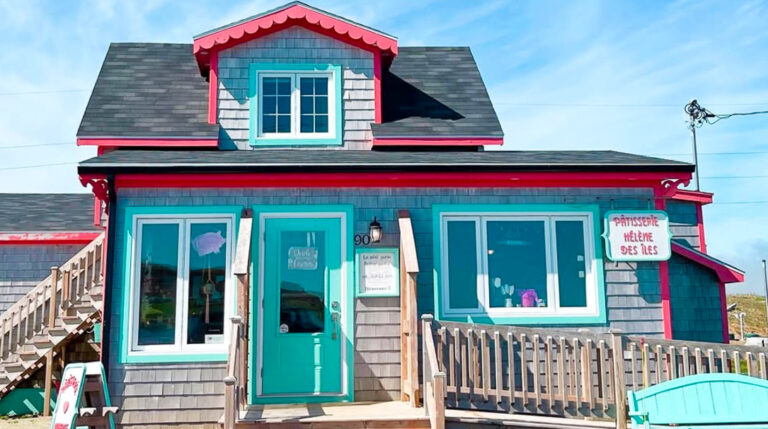 La photo montre la charmante façade de la pâtisserie Hélène des Îles, située aux Îles-de-la-Madeleine. La petite maison, recouverte de bardeaux gris, affiche une allure à la fois rustique et joyeusement colorée. Les moulures et boiseries sont peintes dans des teintes vives de turquoise et de rouge framboise, ce qui lui donne un style accueillant et typiquement madelinot. Une enseigne à droite indique clairement le nom de l’établissement, tandis qu’un écriteau près de la porte turquoise ajoute une touche artisanale. Une rampe de bois encadre l’entrée principale, soulignant l’esprit chaleureux et familial du lieu. Sous un ciel bleu éclatant, cette pâtisserie dégage une atmosphère conviviale qui donne envie d’y entrer pour découvrir ses douceurs.