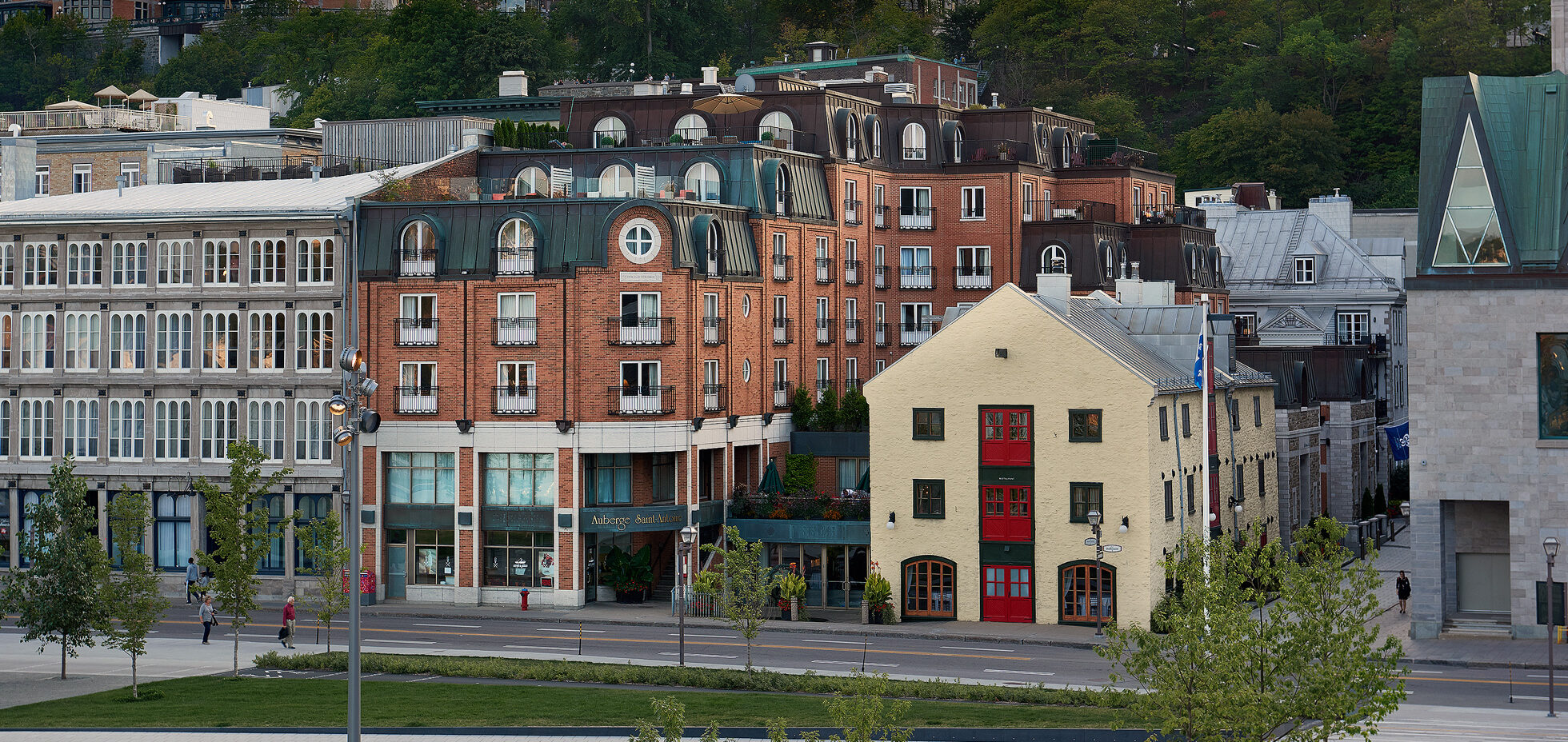 La photo illustre un magnifique panorama urbain du Vieux-Québec, baigné dans une douce lumière de fin de journée. Au premier plan, on distingue des bâtiments emblématiques aux façades variées : à gauche, un édifice gris aux nombreuses fenêtres vitrées; au centre, un immeuble de brique rouge coiffé de toits mansardés verdâtres abritant l’Auberge Saint-Antoine; et à droite, une maison aux murs pâles ponctuée de volets et de portes rouges éclatantes, qui attire immédiatement le regard. En arrière-plan, se dresse le majestueux Château Frontenac, reconnaissable à ses tours et à son imposante silhouette de brique et de cuivre vert-de-gris. Plus loin, on aperçoit aussi l’ancien palais de justice et d’autres édifices historiques de la haute-ville, entourés de verdure. L’ensemble compose un tableau vivant et patrimonial qui exprime toute l’identité architecturale de Québec : un mariage harmonieux entre histoire, charme européen et vitalité contemporaine.