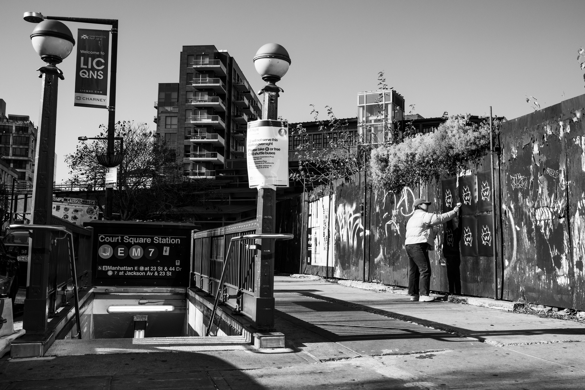 ©Madeleine Dufour. Photo en noir et blanc, prise à Court Square Station (LIC, Queens). À gauche, l’entrée du métro avec les pastilles E / M / 7; lampadaires sphériques, banderole “Welcome to LIC QNS”, tours résidentielles en arrière-plan. À droite, une palissade couverte de graffitis et de peinture écaillée. Une personne en manteau et casquette colle en série des affiches noires ornées d’un même motif blanc répété, évoquant une campagne pour un spiritueux de petit-lait de Charlevoix.