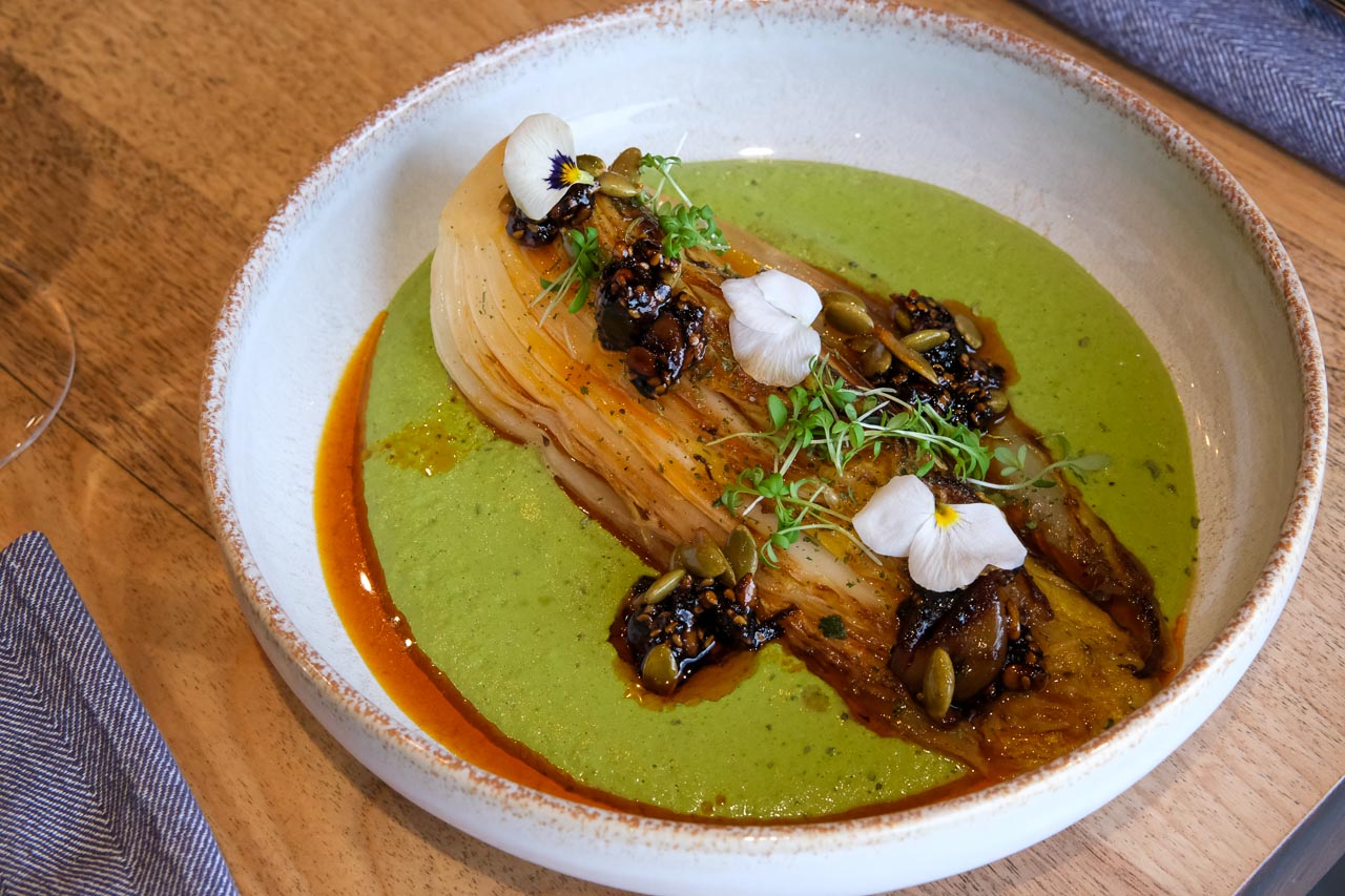 Un chou rôti repose sur une sauce verte lisse et éclatante. un pipián, qui tapisse le fond de l’assiette comme un velours. Autour, un filet d’huile rouge orangé apporte contraste et chaleur visuelle.