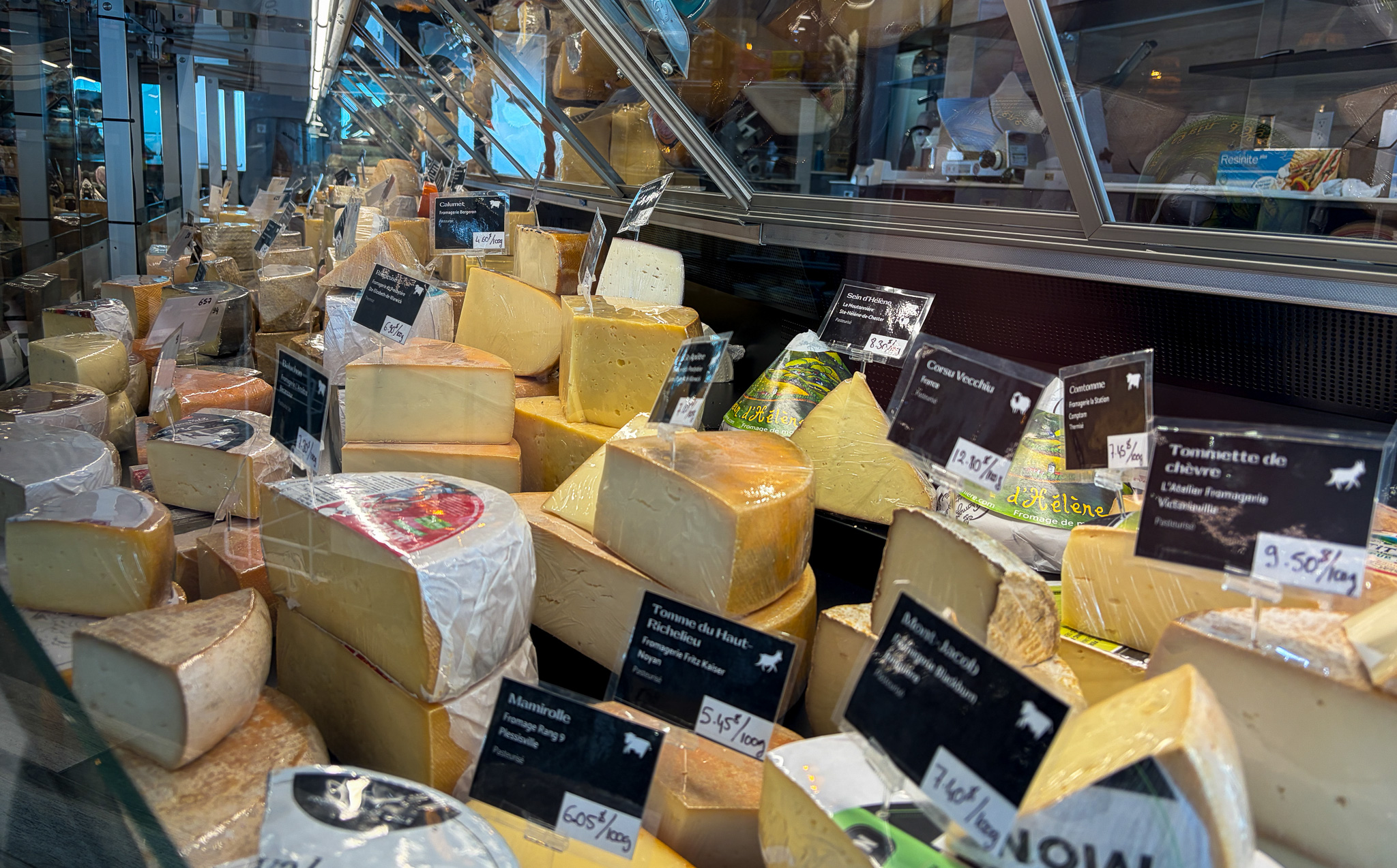 Une vitrine généreusement garnie de fromages occupe tout le cadre. On y voit une grande variété de meules, de pointes et de blocs de fromages québécois et européens, soigneusement disposés derrière la vitre réfrigérée. Les étiquettes noires indiquent les noms, les provenances et les prix, donnant à l’image une impression de comptoir spécialisé, gourmand et vivant. Les textures se répondent : pâtes fermes, croûtes lavées, fromages emballés, tommes, chèvres et fromages affinés. L’ensemble évoque une adresse de quartier où l’on vient autant pour acheter que pour se laisser guider.
