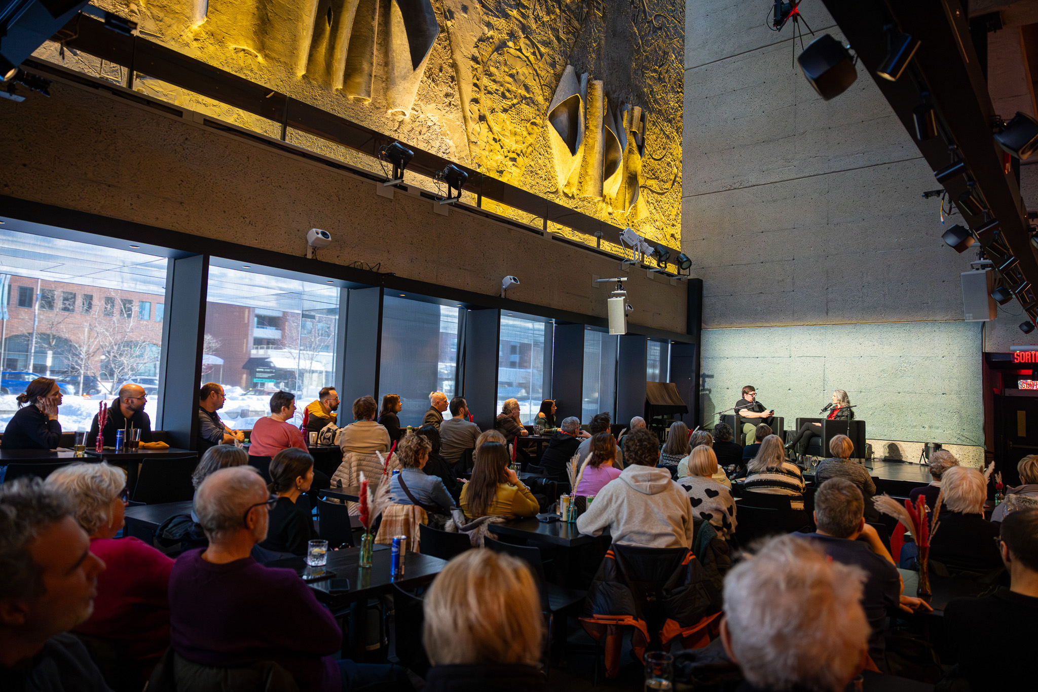 Dans l’espace STUDIOTELUS du Grand Théâtre de Québec, Les rencontres du cœur au ventre prennent vie dans une atmosphère à la fois intime et vibrante. Sous la grande murale baignée de lumière dorée, le public est attablé, attentif, rassemblé autour d’une conversation qui se déroule tout près de lui.