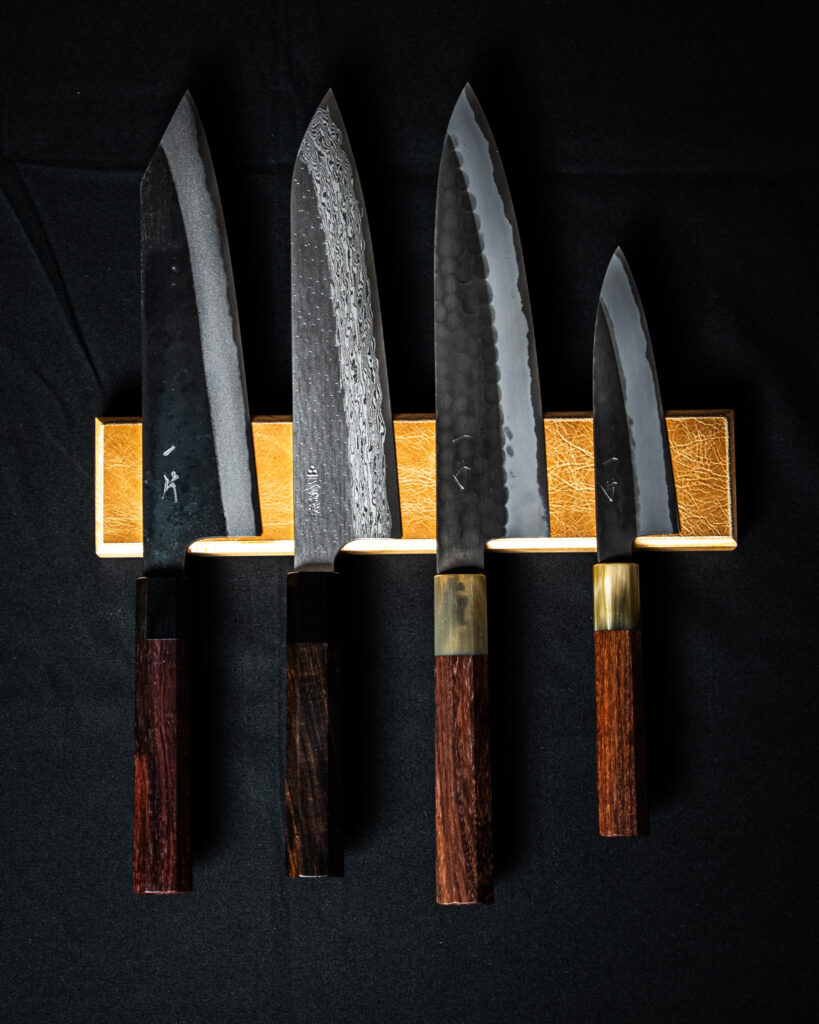 Quatre couteaux japonais aux manches en bois sont présentés sur un support doré devant un fond noir, mettant en valeur les textures des lames et leur finition artisanale.